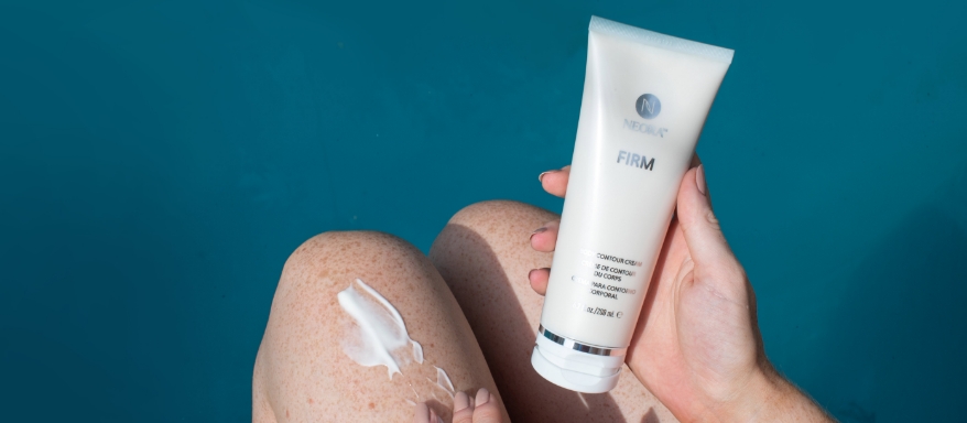 Image of a woman holding a body of Firm Body Contour Cream with product applied to legs with a dark teal background.
